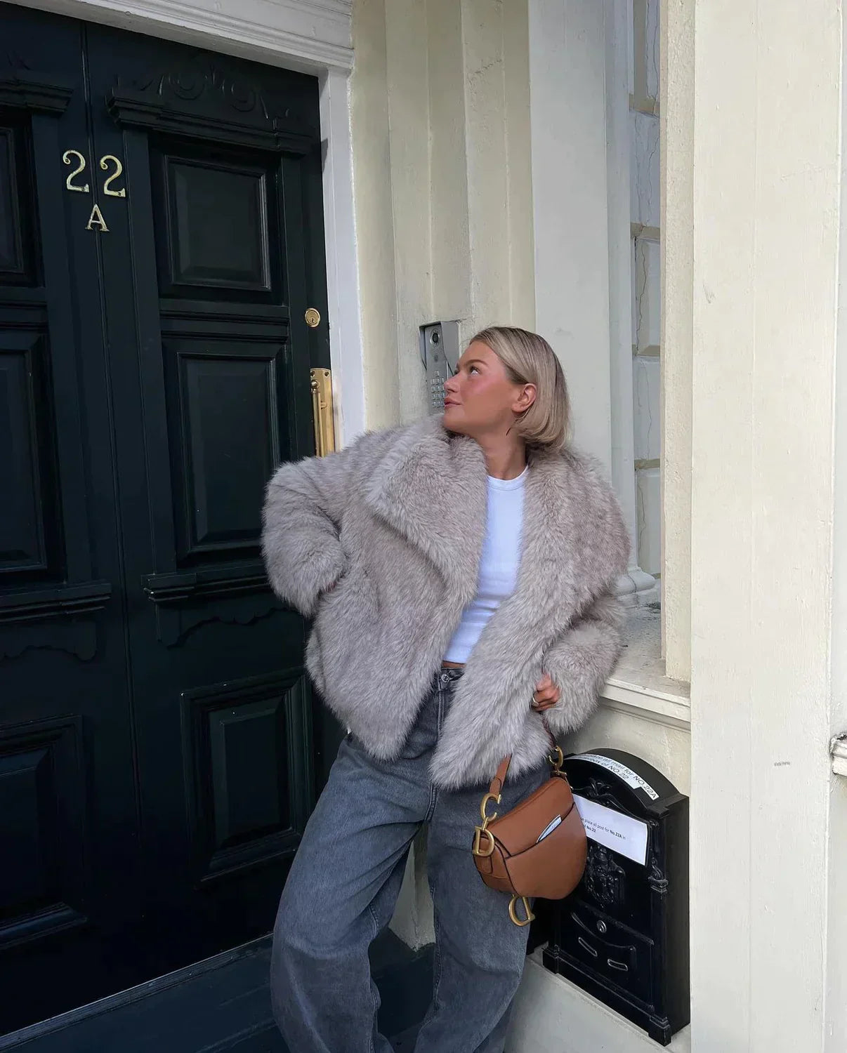 Frau in grauem Kunstpelzmantel vor schwarzer Tür, modisch mit brauner Tasche. Wintermode, Streetstyle, trendige Damenbekleidung.