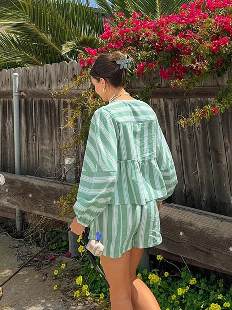 Frau in gestreiftem, grün-weißem Shorts-Anzug, Sommermode, Garten, Blumen im Hintergrund.