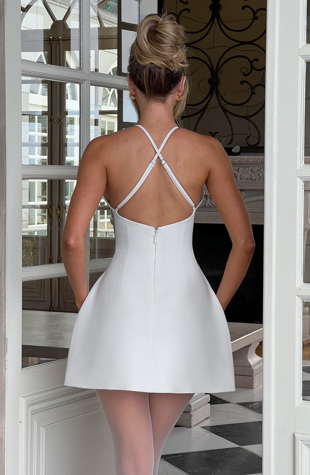 Frau in elegantem, weißem Minikleid mit Kreuzträgern, steht vor einer Tür. Perfekt für Mode, Abendkleider, stilvolle Outfits.