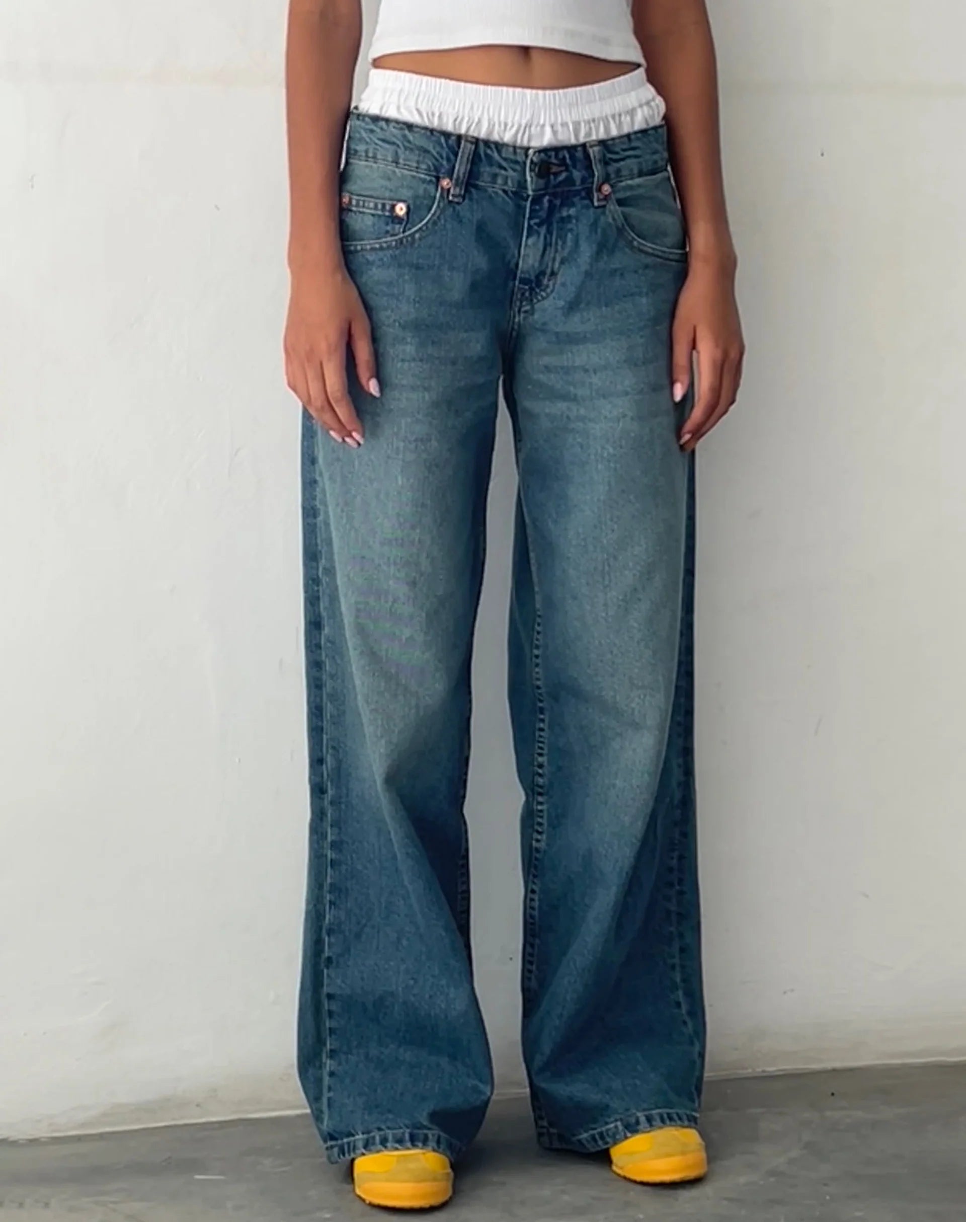 Frau in blauen, weiten Jeans mit gelben Schuhen vor weißer Wand.