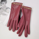 Elegante rote Lederhandschuhe mit goldener Kettenverzierung, stilvoll auf einem Magazin platziert. Perfekt für modische Herbst- und Winteroutfits.