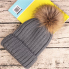 Graue Strickmütze mit Kunstfellbommel auf Holzhintergrund. Wintermode, warme Accessoires, stilvolle Mütze, modische Kopfbedeckung.