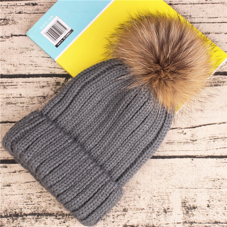 Graue Strickmütze mit Kunstfellbommel auf Holzhintergrund. Wintermode, warme Accessoires, stilvolle Mütze, modische Kopfbedeckung.
