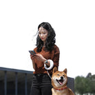Frau mit Smartphone und Shiba Inu Hund an der Leine im Freien, lächelnd, sonniger Tag.