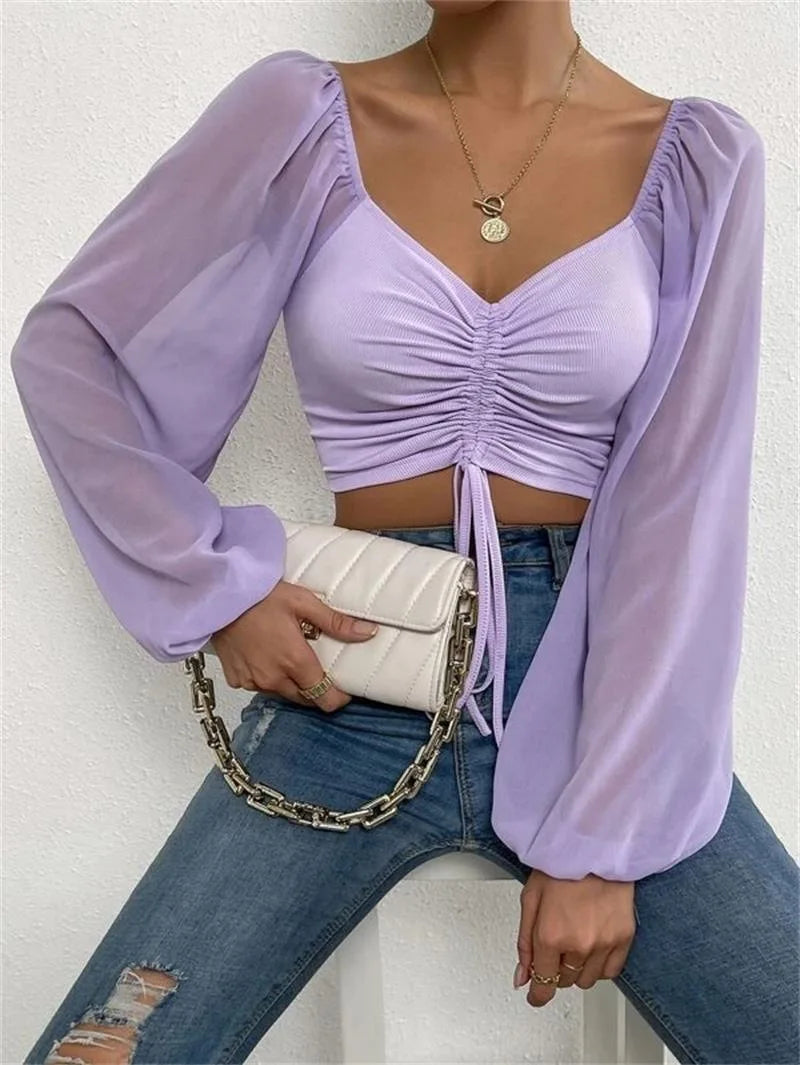 Lila Crop-Top mit langen, transparenten Ärmeln, kombiniert mit zerrissenen Jeans und weißer Kettentasche. Modetrend, Damenmode, Streetstyle.