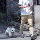 Mann mit weißem Hund an Leine, trägt beige Hose und weißes T-Shirt, hält Kaffeebecher.
