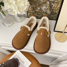 Braune Hausschuhe mit Kunstfellfutter auf weißem Tisch, neben Blumen und Buch. Bequeme, warme Pantoffeln für den Winter. Ideal für Zuhause.