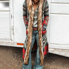 Damenmode: Langer Strickmantel mit Ethno-Muster und Kunstfellbesatz, kombiniert mit Jeans und Gürtel. Perfekt für Herbst und Winter.