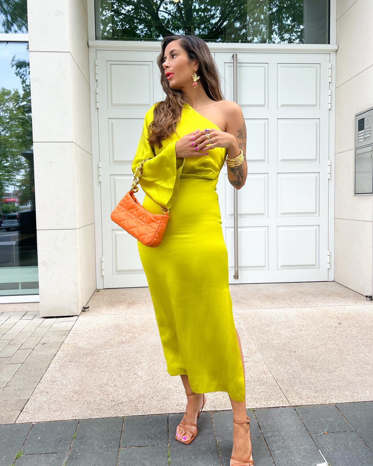 Frau in gelbem Kleid vor weißer Tür, trägt orangefarbene Handtasche. Sommermode, elegantes Outfit, trendige Accessoires, Streetstyle.