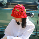 Frau mit rotem, handgestricktem Erdbeer-Hut, sitzt vor einem Auto. Modetrend, handgemachte Accessoires, auffällige Kopfbedeckung.