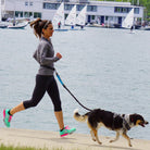 Frau joggt mit Hund am See, Sportbekleidung, Segelboote im Hintergrund, Outdoor-Aktivität.