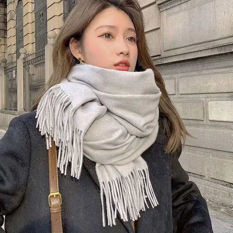 Frau mit grauem Schal und schwarzem Mantel vor städtischer Kulisse. Modetrend, Winteraccessoire, stilvoll, elegant, warm, Damenmode.
