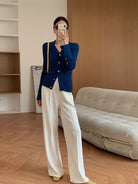 Frau in blauem Strickcardigan und weißen Hosen, steht in modernem Wohnzimmer. Modetrends, Damenmode, eleganter Look, Herbstoutfit.