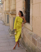 Frau in einem eleganten, einärmeligen gelben Kleid lehnt an einer Steinmauer. Perfekt für Mode, Abendkleider, Streetstyle, Sommermode.