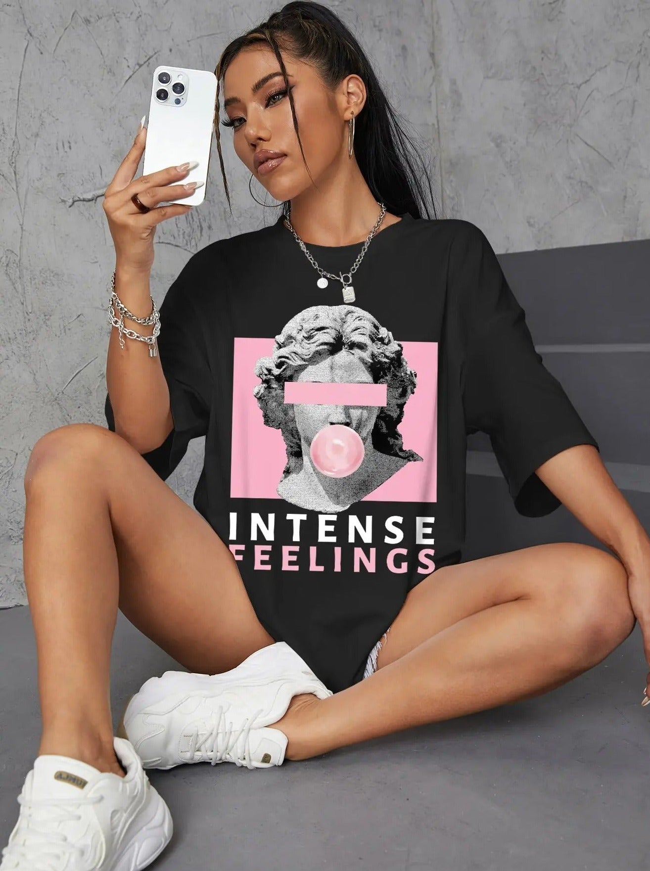Frau in schwarzem T-Shirt mit antikem Skulptur-Design und "Intense Feelings"-Aufdruck, sitzt mit Smartphone, trägt weiße Sneaker.