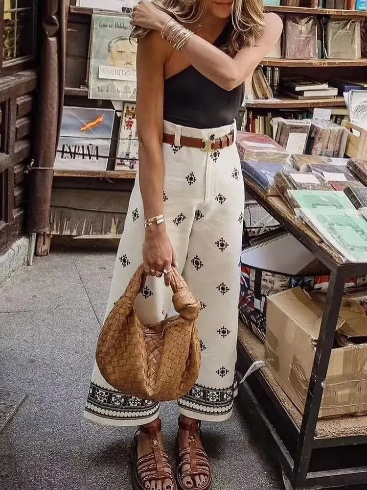 Frau in weißer, bestickter Hose und schwarzem Top, hält eine geflochtene Tasche. Modetrend, Sommeroutfit, Boho-Stil, Streetwear.