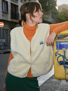 Frau in beiger Fleeceweste und orangefarbenem Rollkragenpullover lehnt an gelbem Briefkasten. Streetstyle, Herbstmode, urbaner Look.