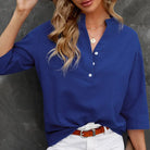 Frau in blauer Bluse mit Knopfleiste, weißer Hose und braunem Gürtel vor grauem Hintergrund.