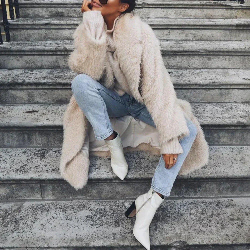 Frau in modischem, flauschigem Kunstpelzmantel, kombiniert mit blauen Jeans und weißen Stiefeletten, sitzt lässig auf Treppe. Wintermode-Trend.