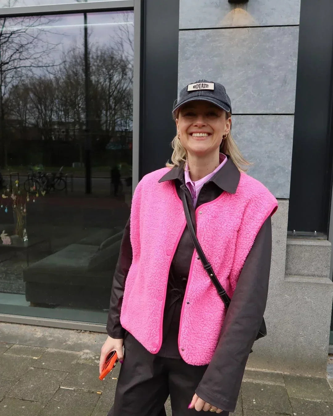 Frau in pinker Weste und schwarzer Kappe vor modernem Gebäude. Streetwear, Mode, urbaner Stil, lässiges Outfit, trendige Kleidung.