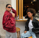 Zwei stilvolle Frauen in modischen Oversize-Jacken vor einem Café. Trendige Streetwear, Sonnenbrillen, lässige Jeans, urbaner Lifestyle.