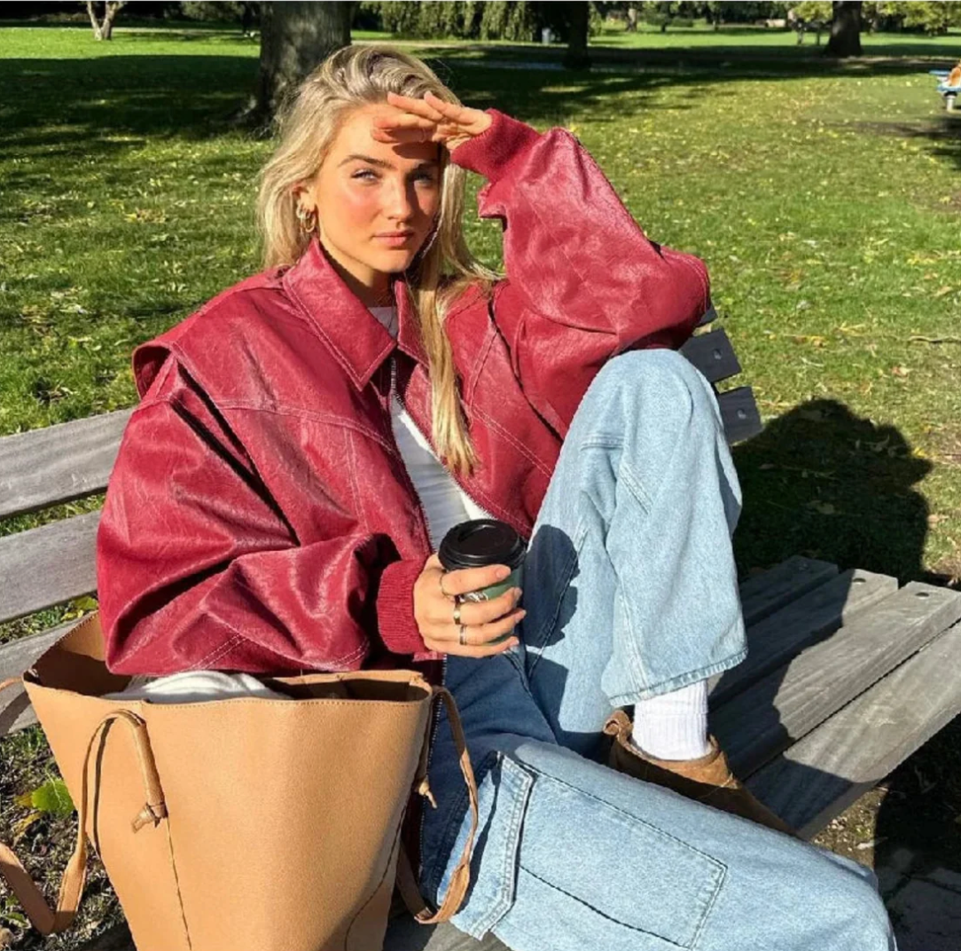 Frau in roter Lederjacke sitzt auf Parkbank, hält Kaffeebecher. Modetrend Herbst, lässiger Stil, Outdoor-Mode, Damenbekleidung.