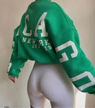 Grüner Oversize-Pullover mit weißem "New York" Print, kombiniert mit weißen, gerippten Radlerhosen. Trendige Streetwear-Mode für Damen.