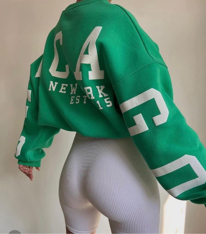 Grüner Oversize-Pullover mit weißem "New York" Print, kombiniert mit weißen, gerippten Radlerhosen. Trendige Streetwear-Mode für Damen.