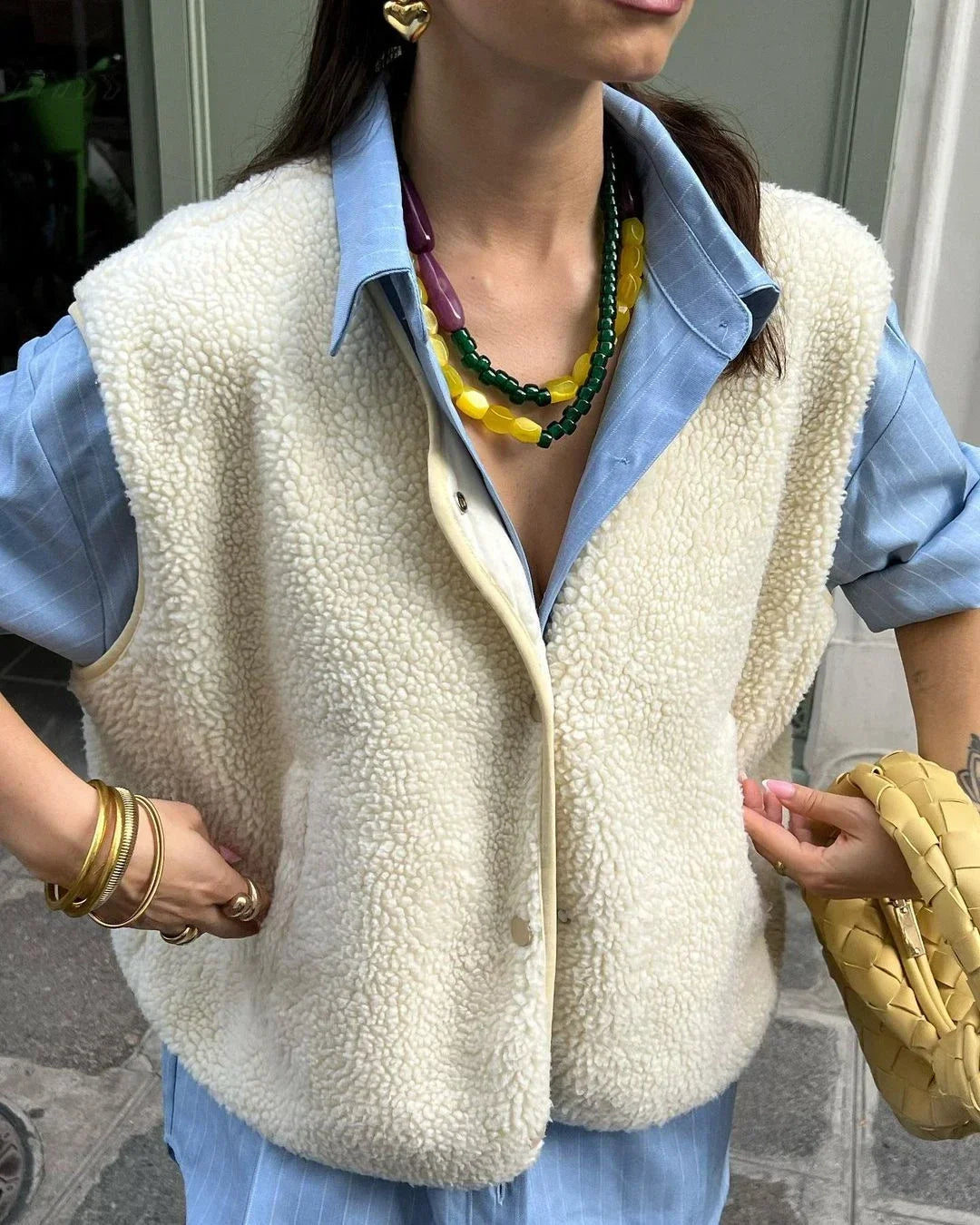 Frau trägt modische Schafwollweste über blauem Hemd, kombiniert mit bunten Halsketten und goldenen Armreifen. Trendige Herbstmode 2023.