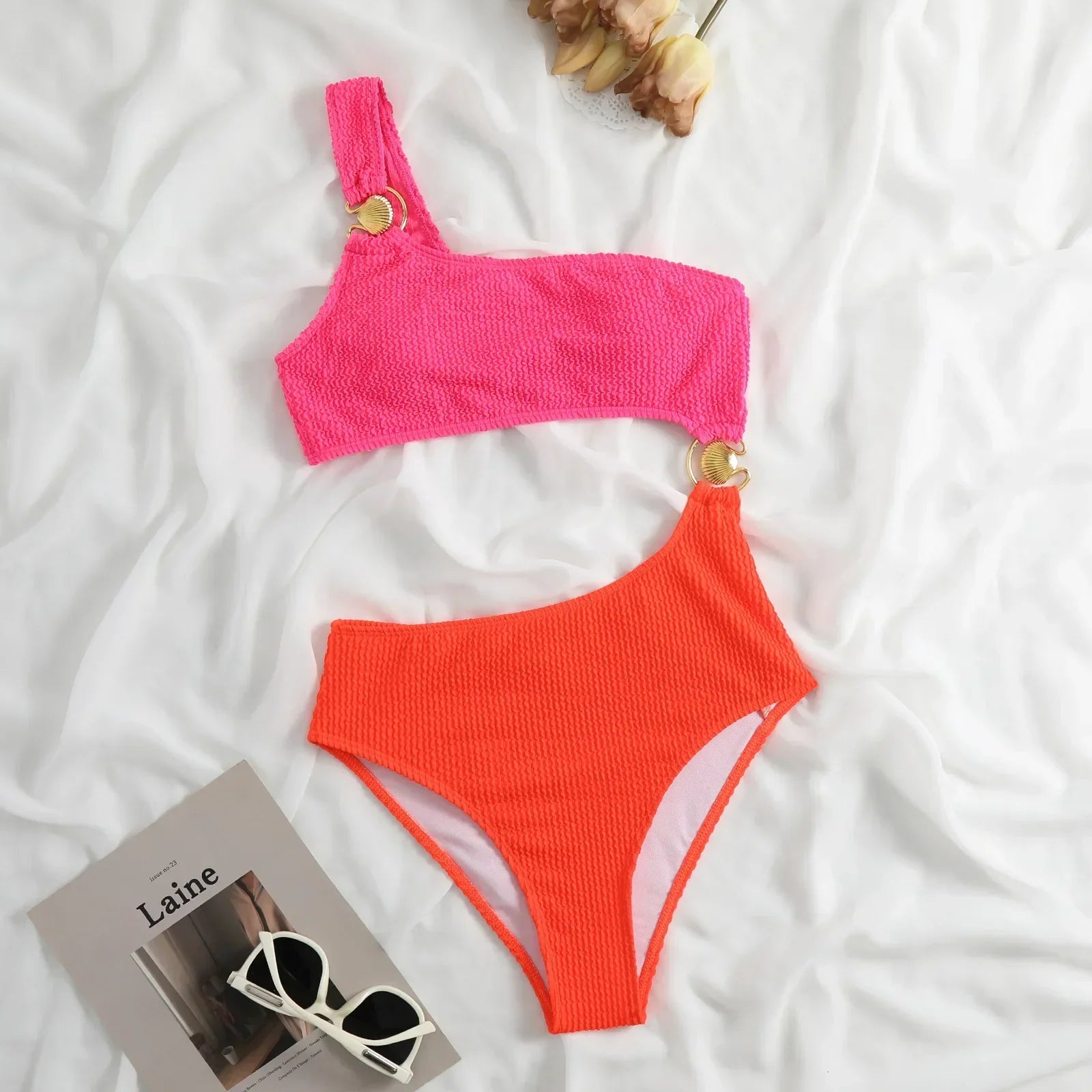 Neonfarbener Bikini mit asymmetrischem Design, goldenen Ringen, auf weißem Hintergrund.