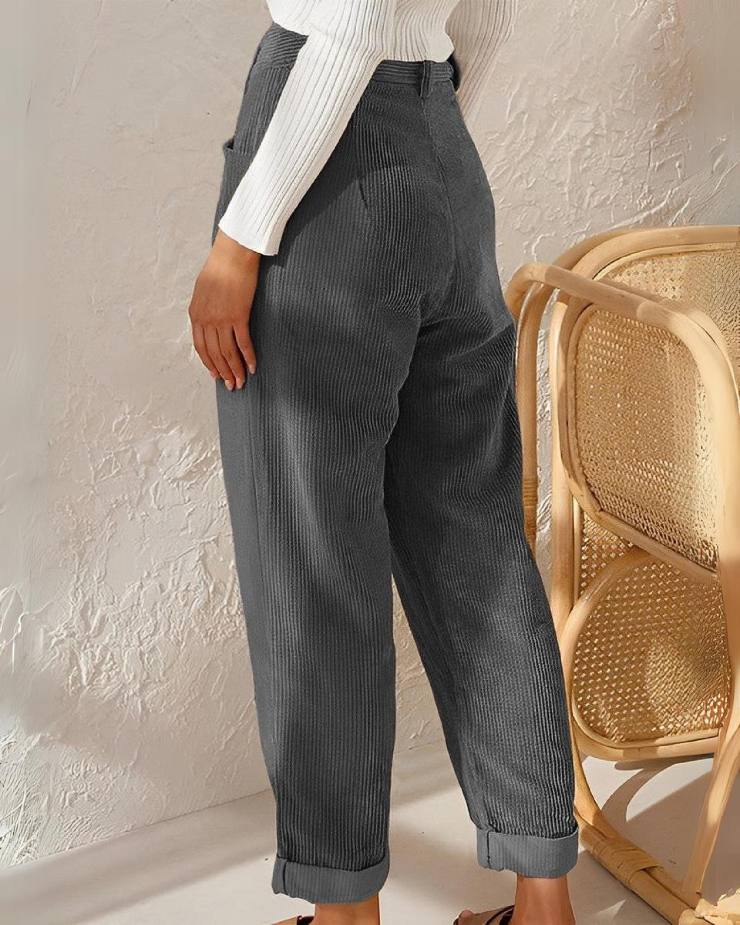 Frau in grauer Cordhose, weißem Pullover, neben Rattan-Stuhl, modisch, lässig, Herbstmode.