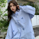 Frau in hellblauer Oversize-Bluse mit Schmetterlingsstickerei, modisch und lässig, ideal für trendige Streetwear-Looks.