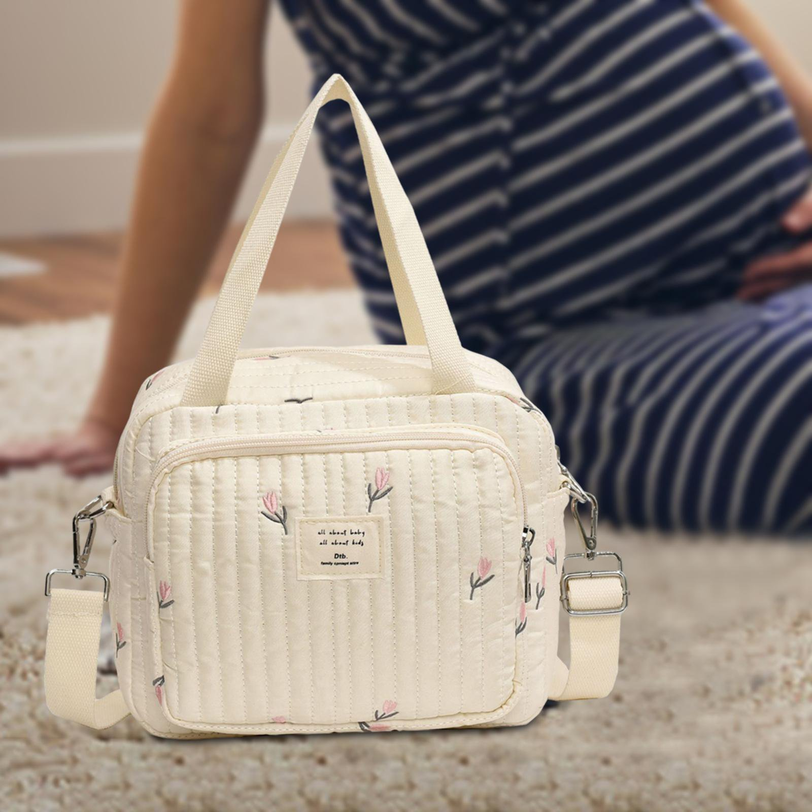 Weiße Wickeltasche mit Blumenmuster, gesteppt, vor schwangerer Frau auf Teppich.