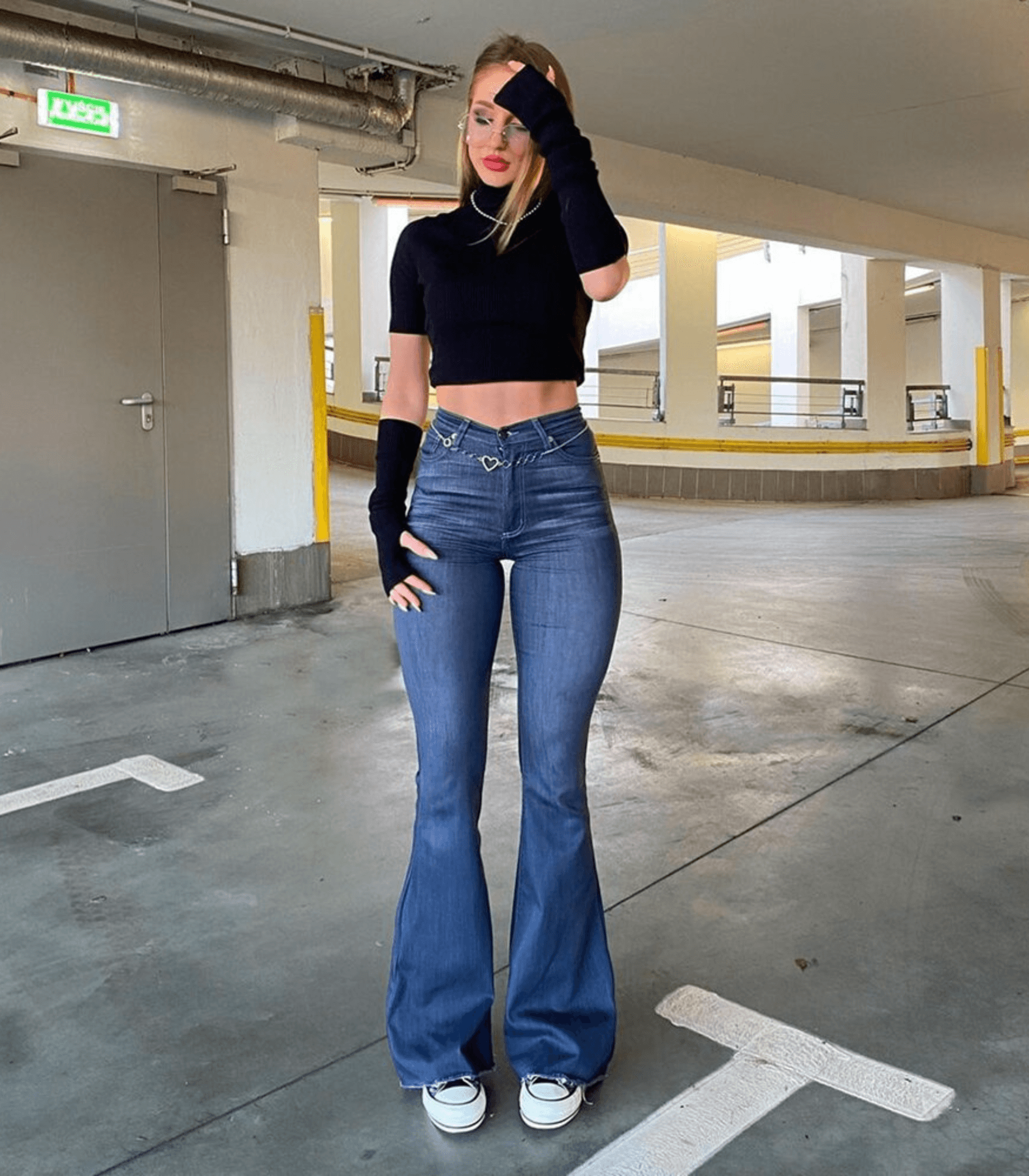 Frau in hoch taillierter, ausgestellter Stretch-Jeans, kombiniert mit schwarzem Crop-Top und Sneakers. Modetrend, bequeme Passform, urbaner Stil.
