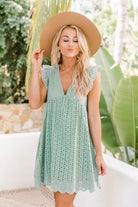 Frau in grünem Sommerkleid mit Spitzenmuster und Strohhut, stehend vor tropischem Hintergrund. Perfekt für Sommermode und Strandoutfits.