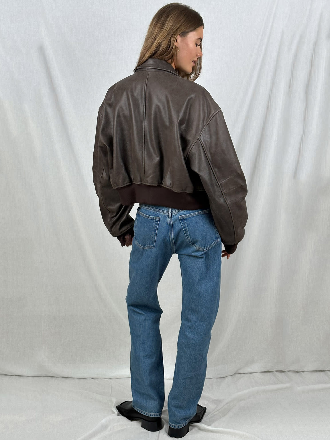 Frau in brauner Lederjacke und blauer Jeans, Rückansicht. Modetrend, lässiger Stil, hochwertige Damenbekleidung, zeitloses Design.