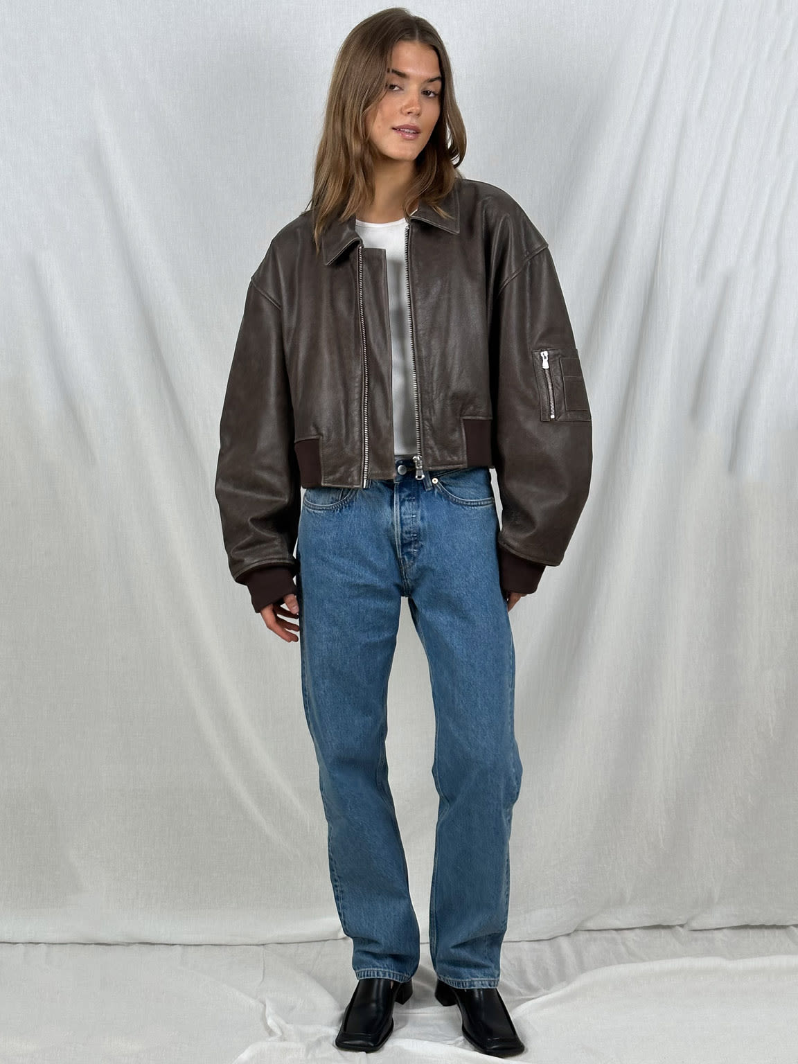 Frau in brauner Lederjacke, blauer Jeans und schwarzen Stiefeln vor weißem Hintergrund. Modetrend, lässiger Stil, Damenbekleidung.