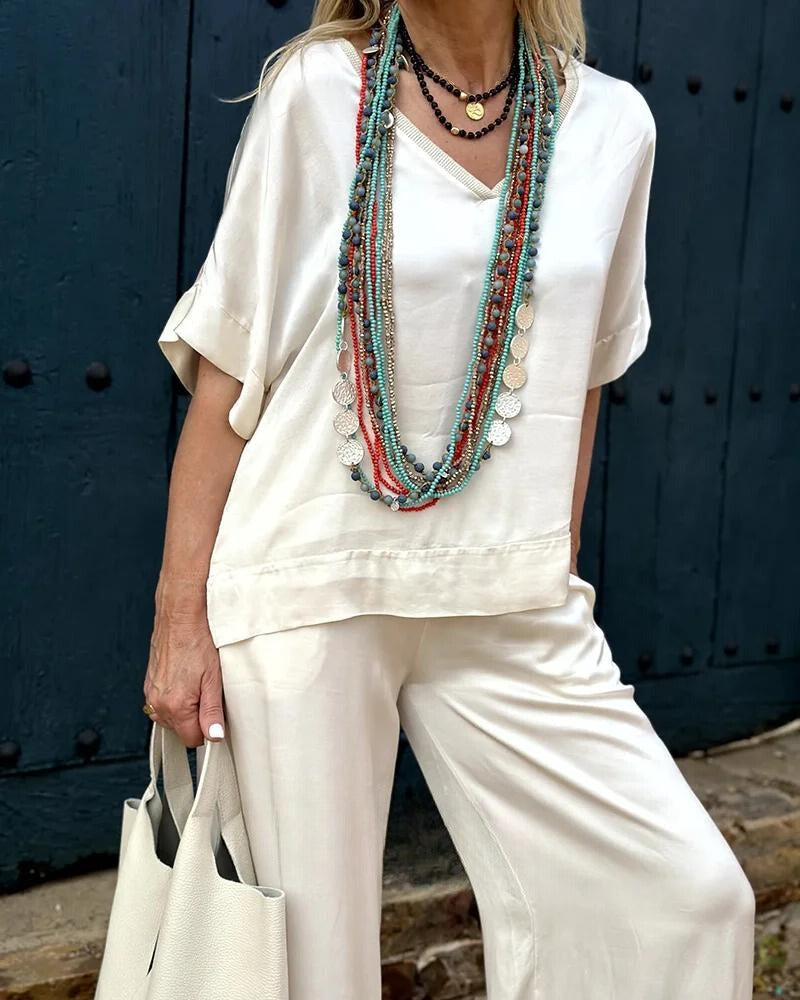 Frau in eleganter, weißer Leinenbluse und Hose, trägt bunte Statement-Ketten. Sommermode, Damenbekleidung, lässiger Stil, Accessoires.
