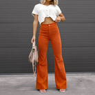 Frau in weißem Strickoberteil und orangefarbener Schlaghose vor grauer Wand, hält beigefarbene Handtasche. Modetrend, Damenmode, Herbstoutfit.