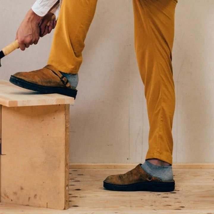 Person trägt braune Clogs und gelbe Hose, steht auf Holzplattform. Modische Herrenschuhe, handgefertigt, bequem, ideal für Freizeit und Alltag.