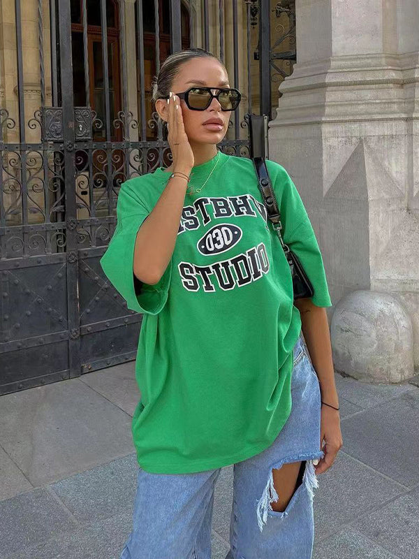 Frau in grünem Oversize-T-Shirt mit "ESTHER STUDIO"-Aufdruck, zerrissene Jeans, Sonnenbrille; urbaner Streetstyle, modisch, lässig.