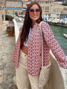 Frau in modischer Jacke mit geometrischem Muster, lässiger weißer Hose und gehäkelter Tasche posiert lächelnd am Hafen. Trendige Damenmode.