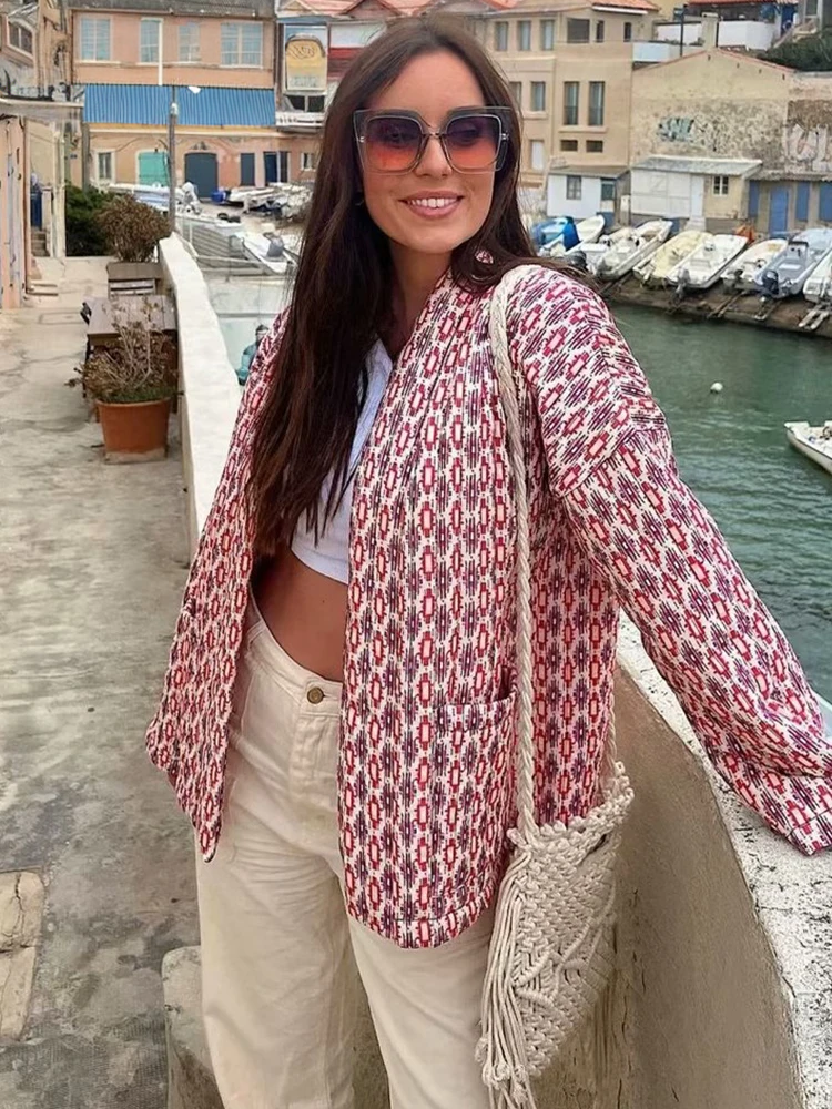 Frau in modischer Jacke mit geometrischem Muster, lässiger weißer Hose und gehäkelter Tasche posiert lächelnd am Hafen. Trendige Damenmode.