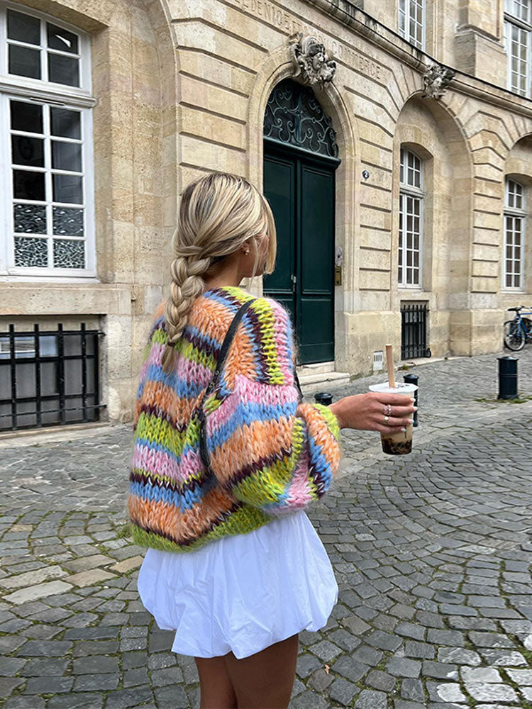 Frau in bunter Strickjacke und weißem Kleid hält Bubble Tea. Modetrend, Streetstyle, Herbstmode, Strickmode, Stadtspaziergang.