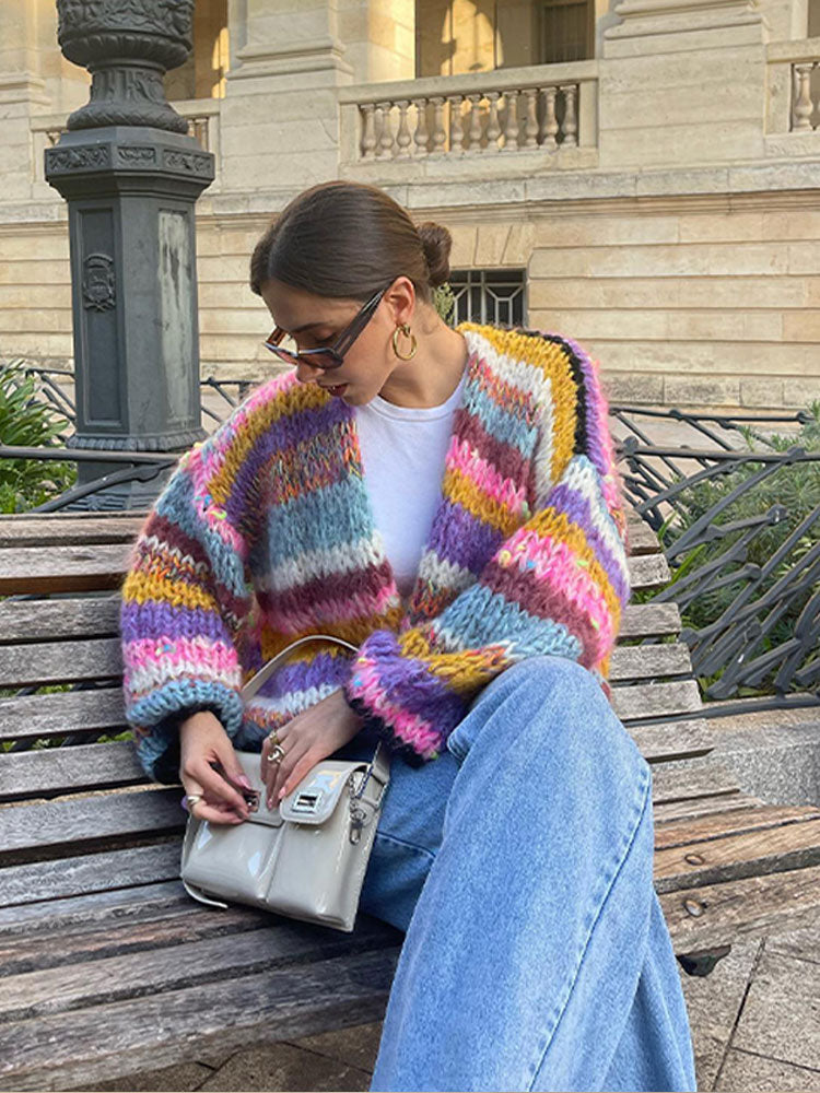Frau mit buntem Strickcardigan und blauer Jeans sitzt auf einer Bank. Modetrend, Herbstmode, Strickjacke, lässiger Stil, Damenmode.