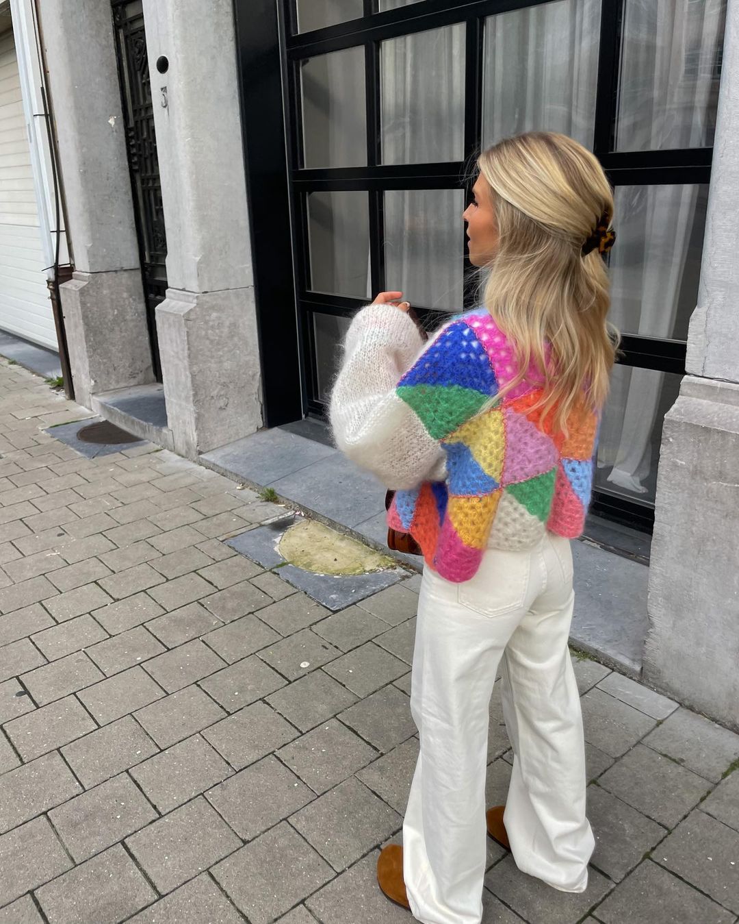 Frau in bunter Strickjacke mit geometrischem Muster, weiße Hose, braune Schuhe. Modetrend, Streetstyle, Herbstmode, Damenbekleidung.