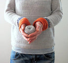 Grauer Boho-Pullover mit orangefarbenen Bündchen, handgehaltenes Garnknäuel, lässiger Stil, ideal für Herbstmode und DIY-Projekte.