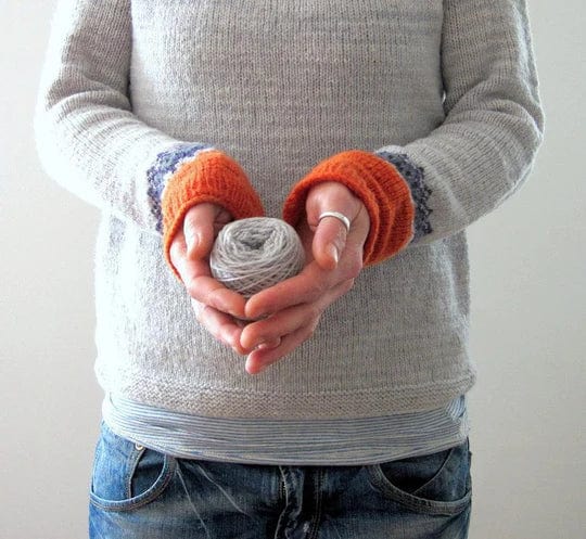 Grauer Boho-Pullover mit orangefarbenen Bündchen, handgehaltenes Garnknäuel, lässiger Stil, ideal für Herbstmode und DIY-Projekte.