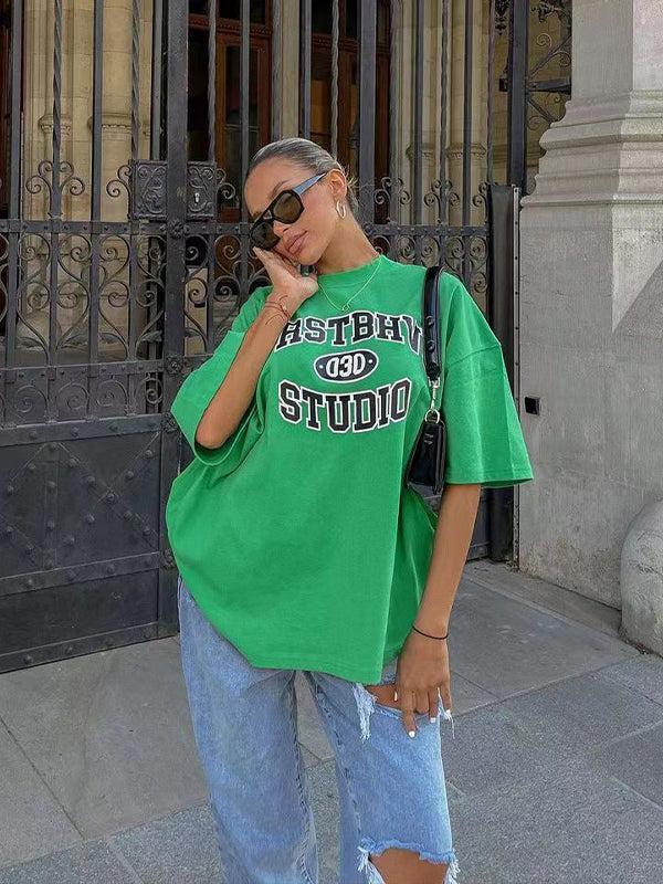 Frau in grünem Oversize-T-Shirt mit "Astrow Studio"-Aufdruck, kombiniert mit blauer Jeans und Sonnenbrille, posiert vor einem historischen Gebäude.
