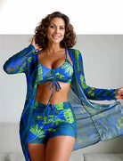 Frau in tropischem, blau-grünem Strandoutfit mit Blättermuster, bestehend aus Bikini, Shorts und transparentem Kimono. Sommermode, Strandkleidung.